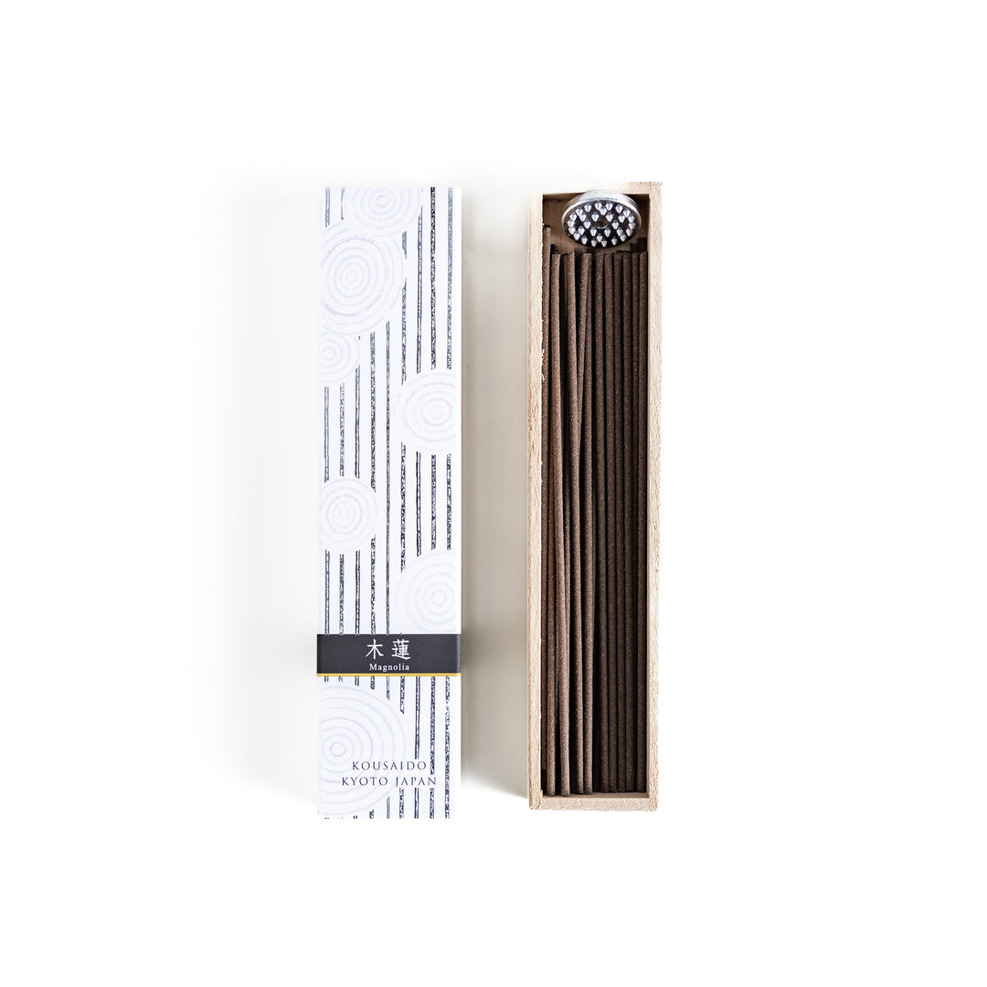 Incense in a light wooden box with deep brown sticks, elegantly packaged in a white design with subtle circular patterns and black accents, evoking a refined magnolia scent.