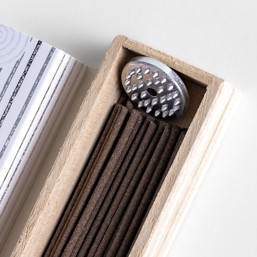 Incense in a light wooden box with deep brown sticks, elegantly packaged in a white design with subtle circular patterns and black accents, evoking a refined magnolia scent.