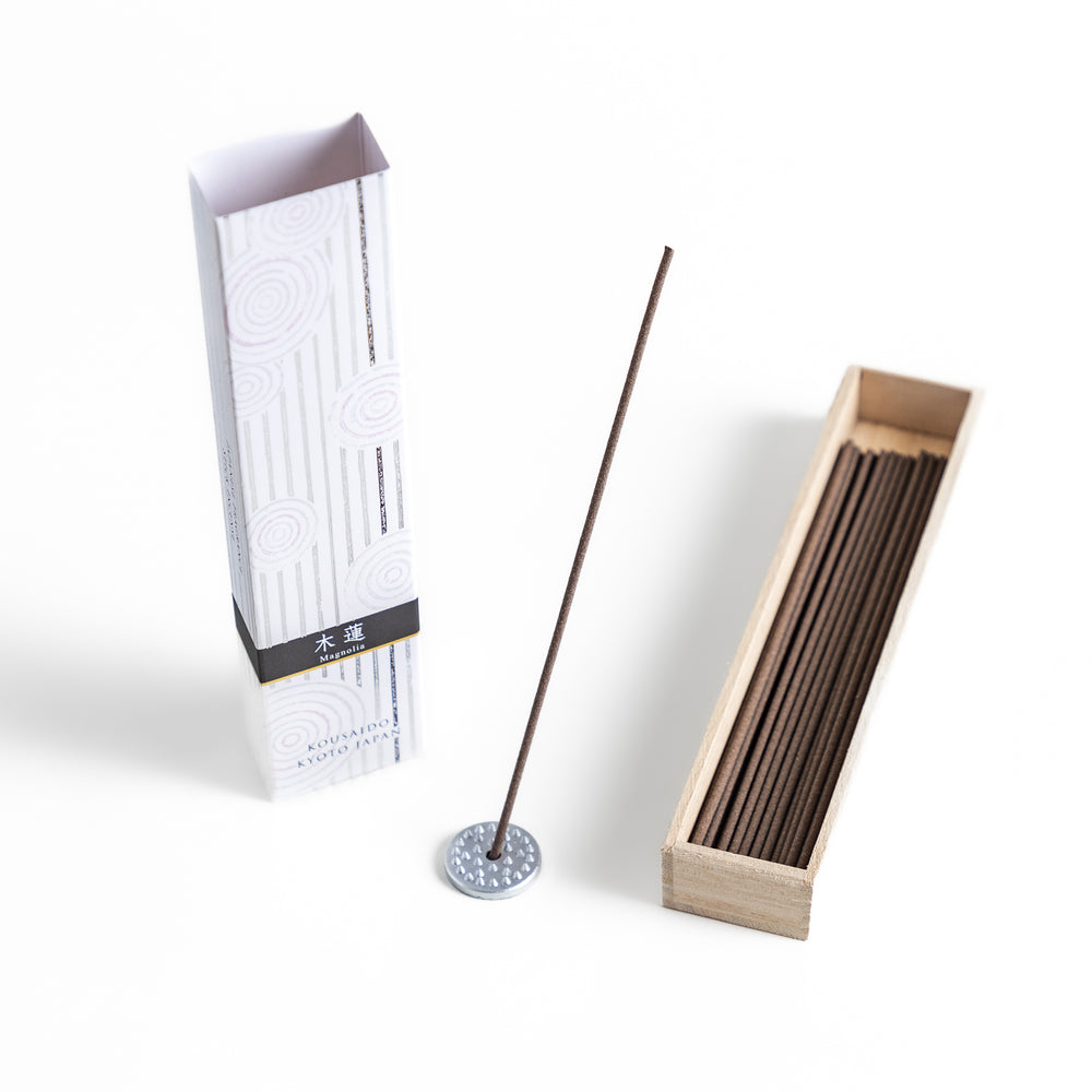 Incense in a light wooden box with deep brown sticks, elegantly packaged in a white design with subtle circular patterns and black accents, evoking a refined magnolia scent.