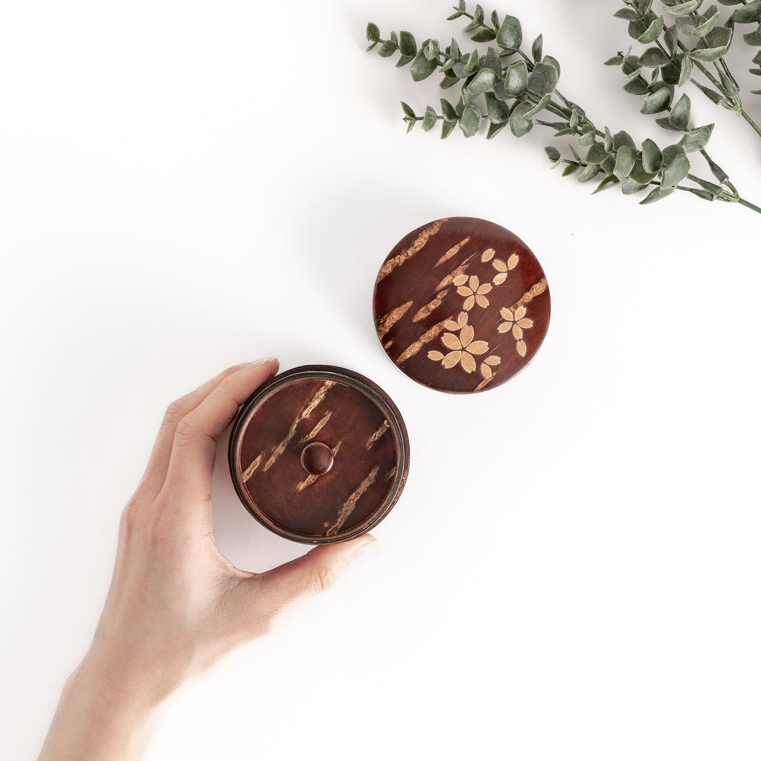 Hand holding a cherry bark tea canister with a lid, showcasing its rich brown color and delicate cherry blossom inlay, perfect for storing tea.