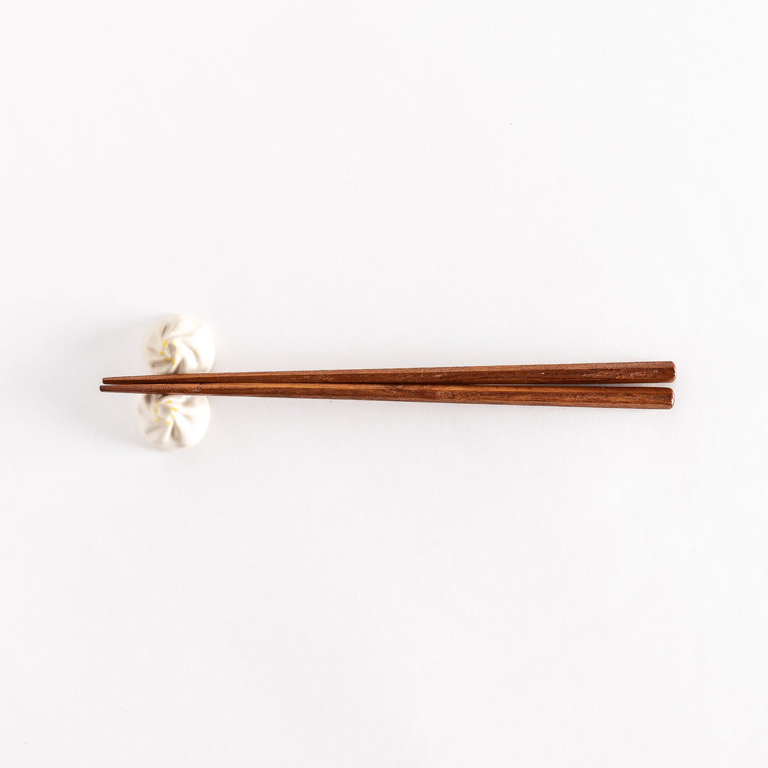 Wide shot of wooden chopsticks resting on a soup dumpling-shaped chopstick rest, placed on a white surface.