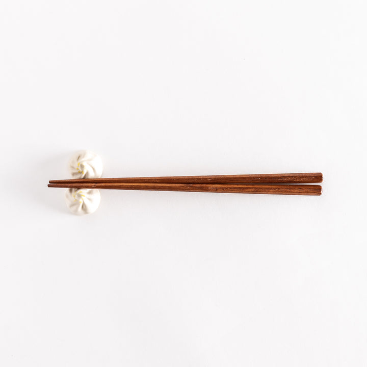 Wide shot of wooden chopsticks resting on a soup dumpling-shaped chopstick rest, placed on a white surface.