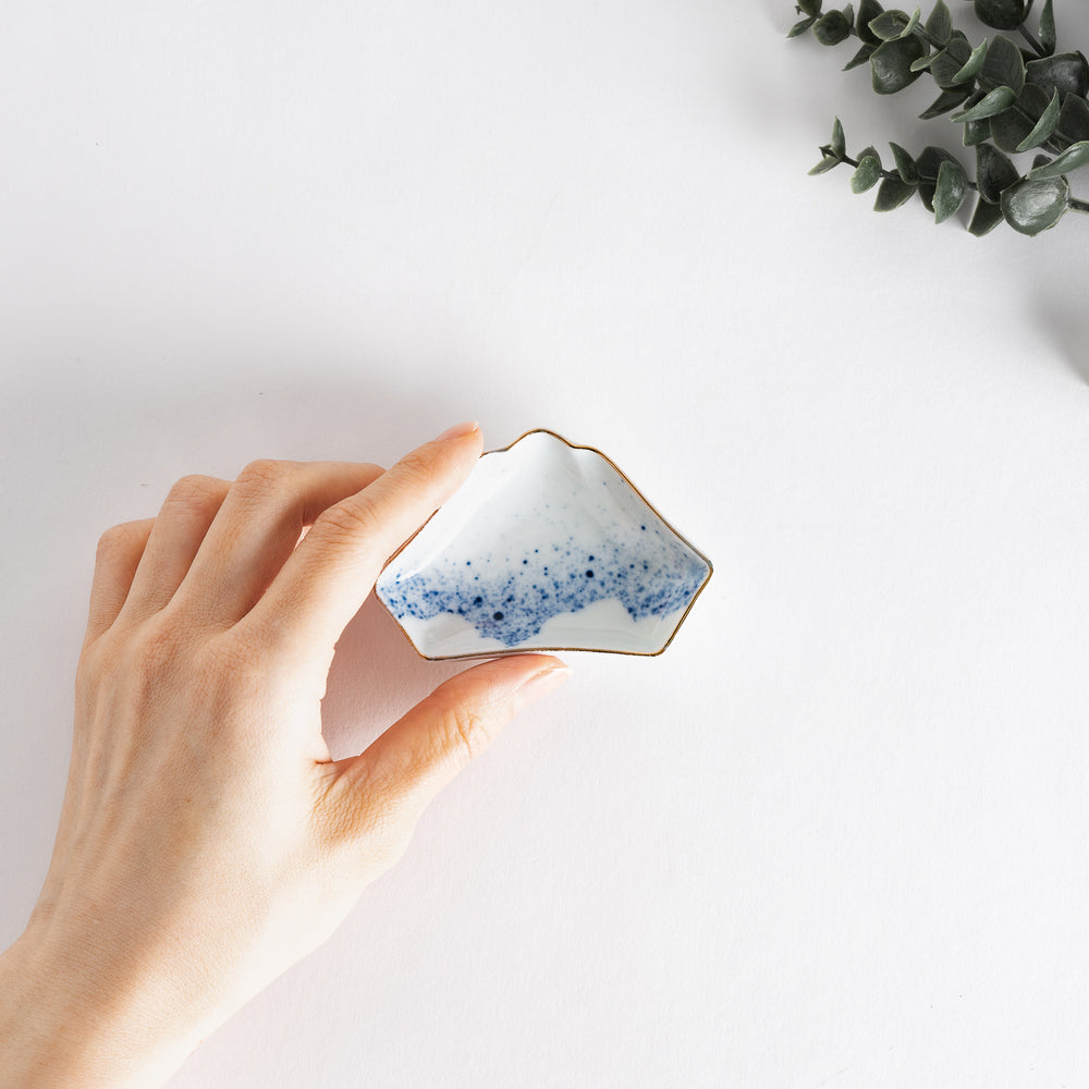 A hand holding a small mountain-shaped sauce dish with a sprayed ink design in blue at the base, featuring a delicate gold rim and elegant shape.