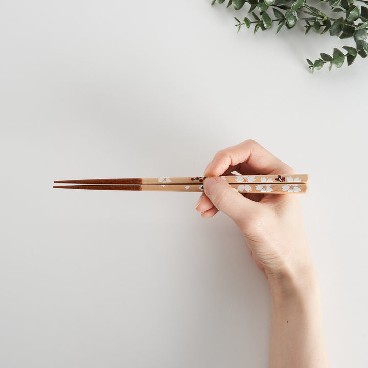 A hand gracefully holds chopsticks with a delicate floral design, featuring white flowers with a red accent.