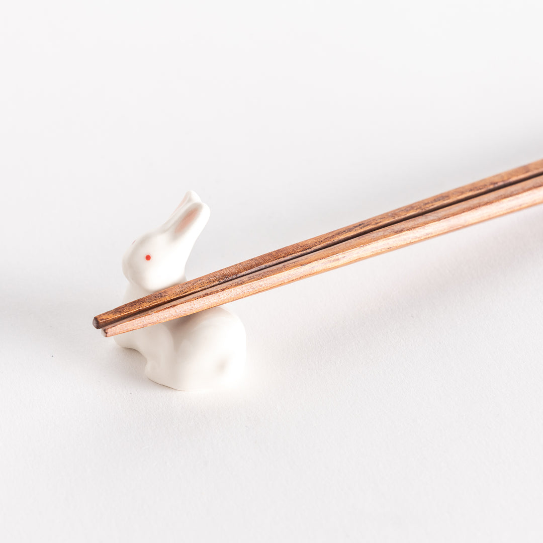 A close-up of a ceramic white rabbit chopstick rest holding a pair of chopsticks, highlighting its delicate design and red eyes.