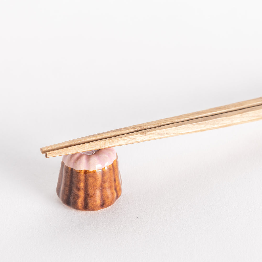 A close-up of a strawberry canelé-shaped ceramic chopstick rest holding chopsticks, highlighting its glossy pink top and detailed texture.