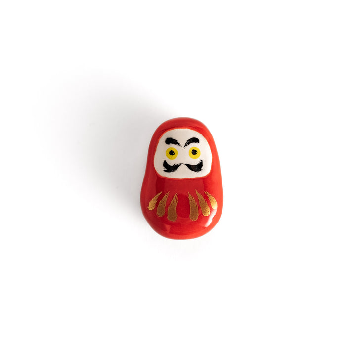 A red daruma doll-shaped chopstick rest with bold black and yellow facial features and gold accents on a white background.