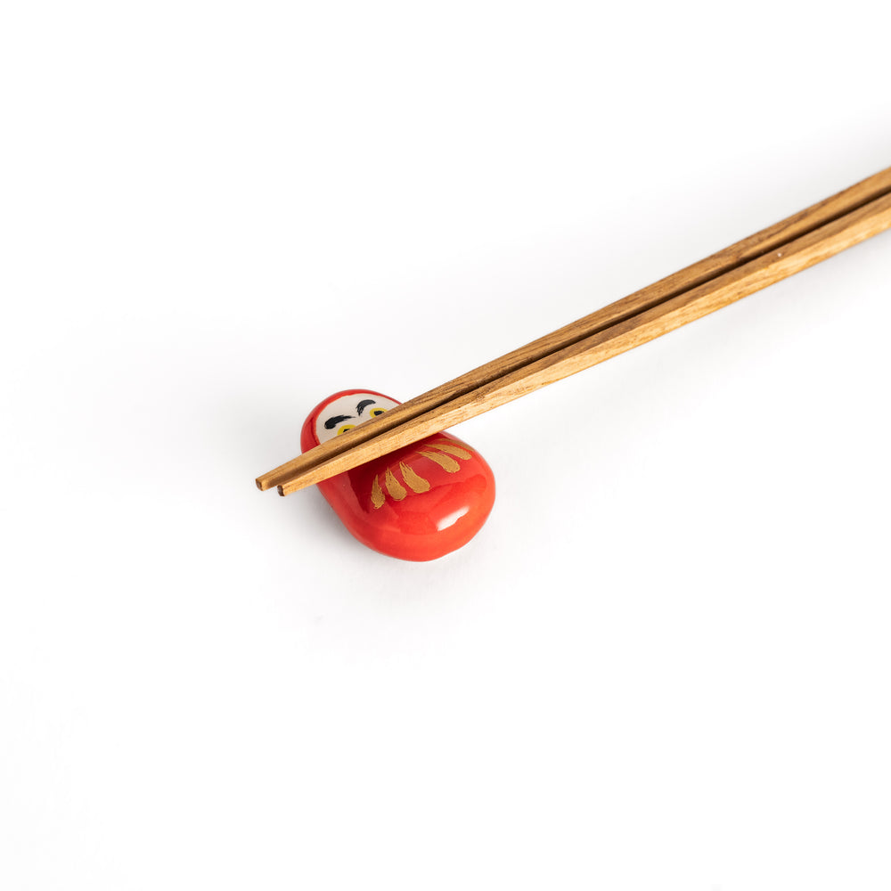 A close-up view of chopsticks resting on the red daruma chopstick rest, highlighting its intricate gold and facial details.