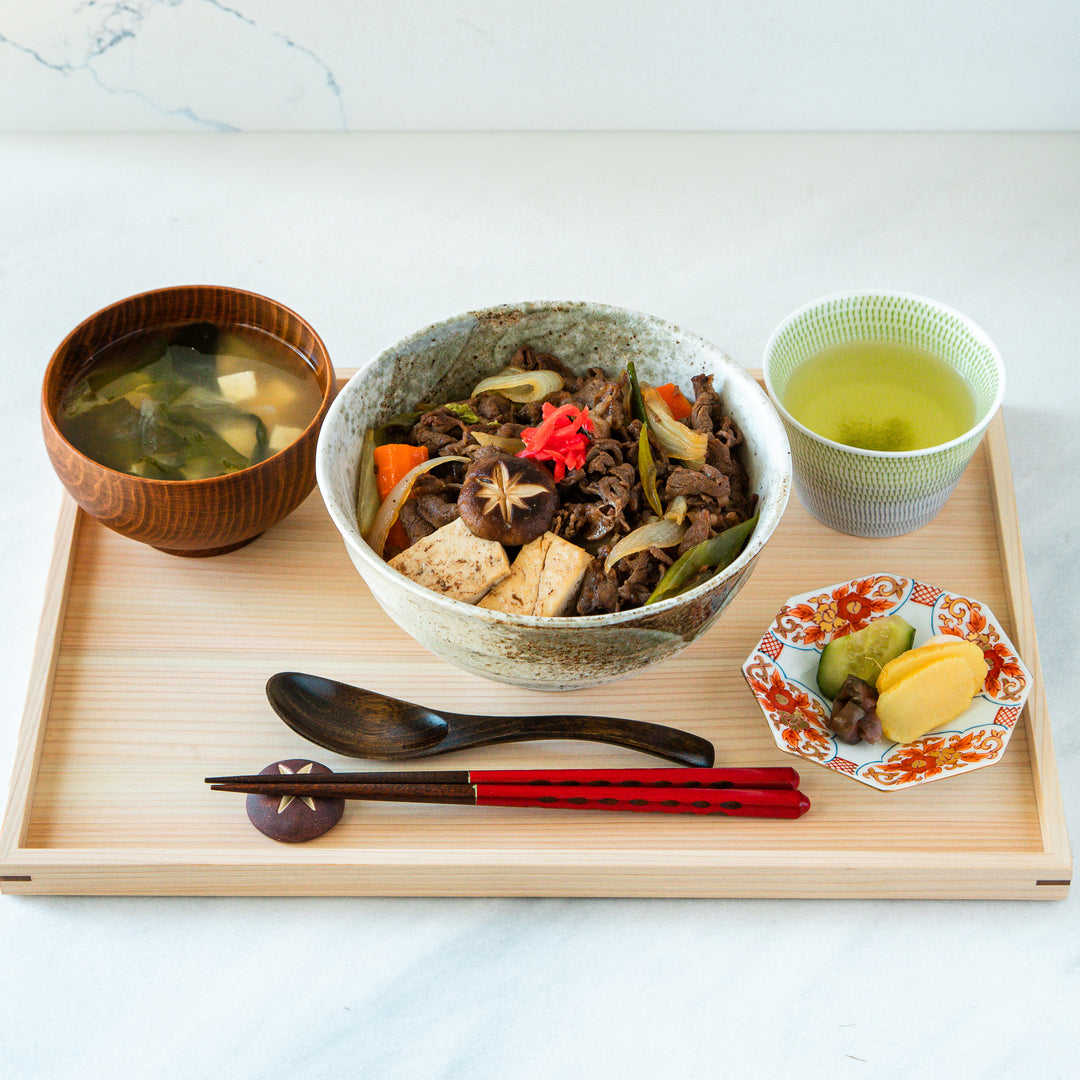 Top view of a sukiyaki donburi bowl with beef, tofu, vegetables, and pickled ginger, served with miso soup, green tea, and pickled vegetables on the side.