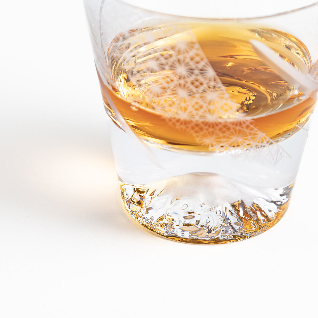 Close-up of a glass with a geometric design, showing whisky and Mount Fuji base in the clear, with detailed etched patterns.





