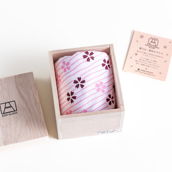 A sakura-themed glass wrapped in a pink and white striped cloth with flower patterns, placed in a wooden box, accompanied by a product card.