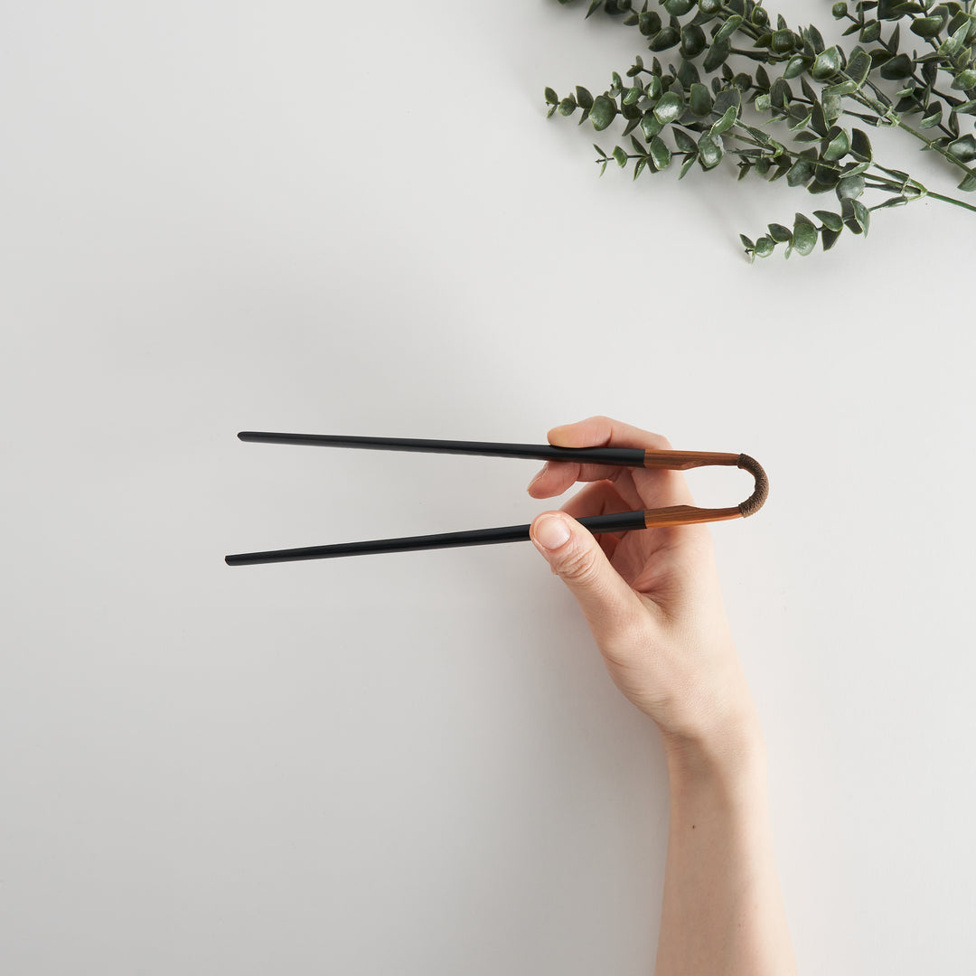 Hand holding black lacquered chopsticks with a curved wooden handle, showing the twine wrapping.