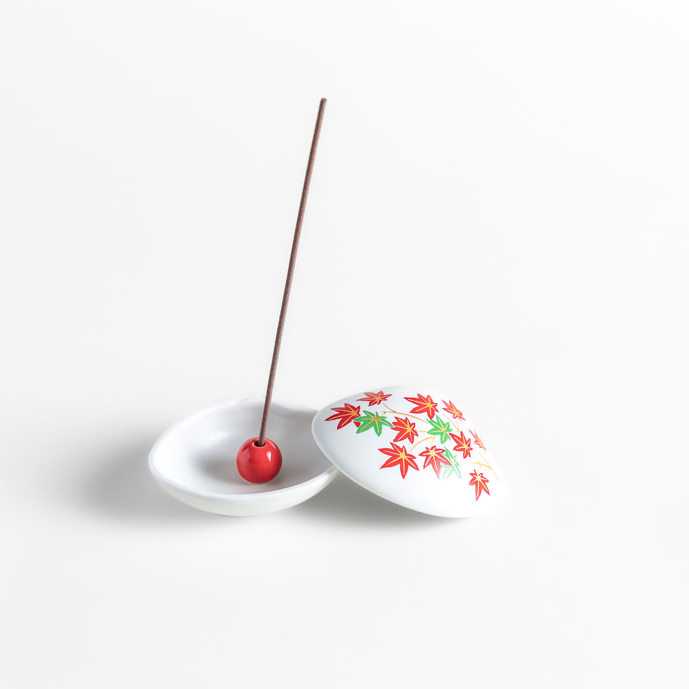 White ceramic incense holder shaped like a seashell, adorned with red and green maple leaves, featuring a small red incense stand inside for secure placement.