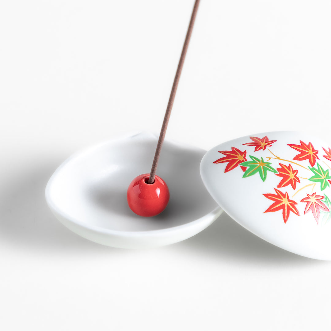 White ceramic incense holder shaped like a seashell, adorned with red and green maple leaves, featuring a small red incense stand inside for secure placement.