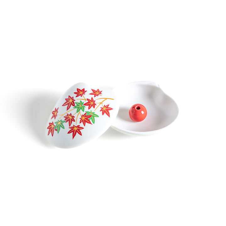 White ceramic incense holder shaped like a seashell, adorned with red and green maple leaves, featuring a small red incense stand inside for secure placement.