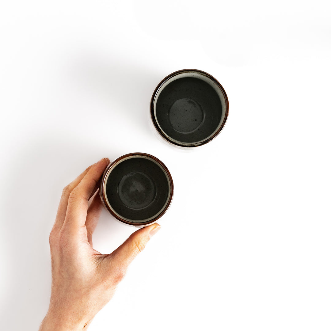 Hand holding a small teacup with a dark glaze, next to a similar teacup, viewed from above.