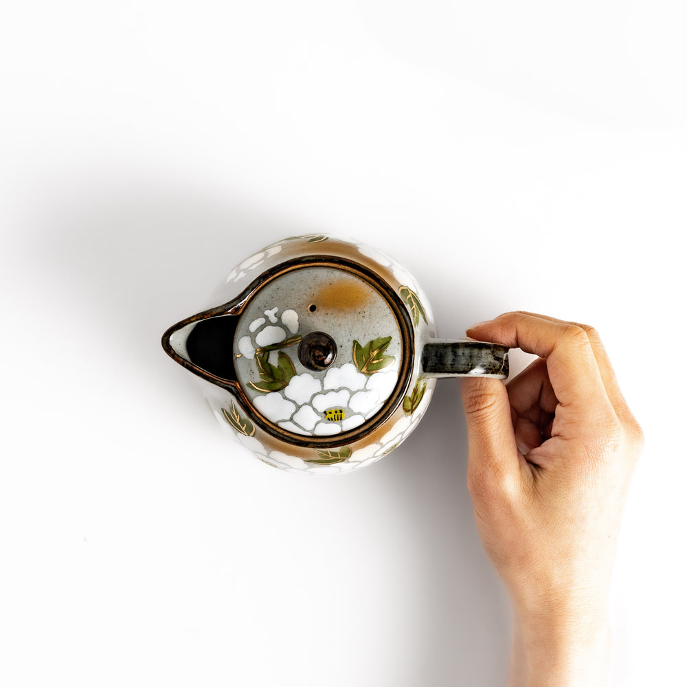 Top-down view of the teapot with white peony patterns, gold highlights, and a hand holding the handle, showcasing its detail and charm.