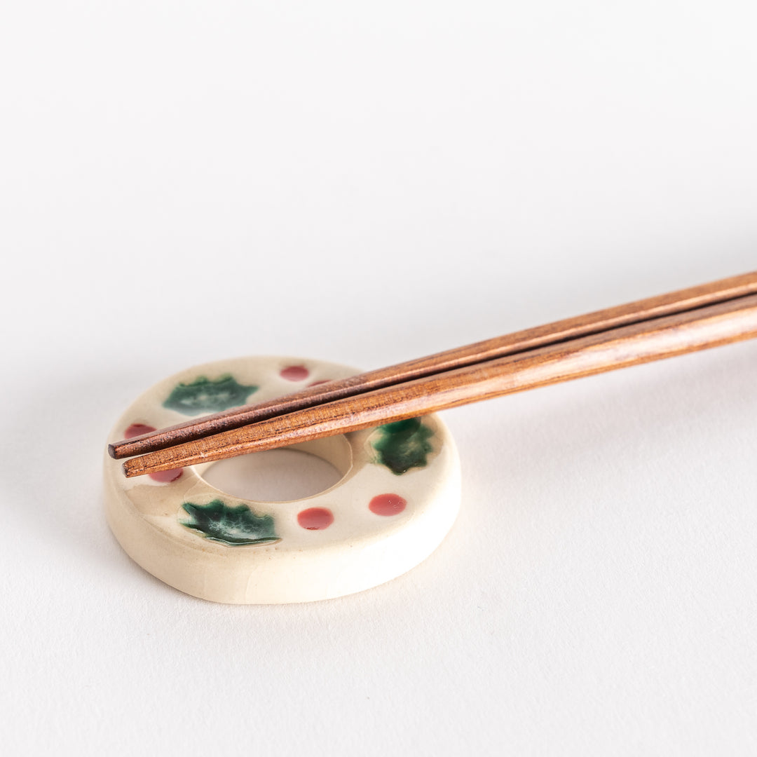 A close-up of a white wreath-shaped chopstick rest with chopsticks placed on it, showing its green and red festive design.