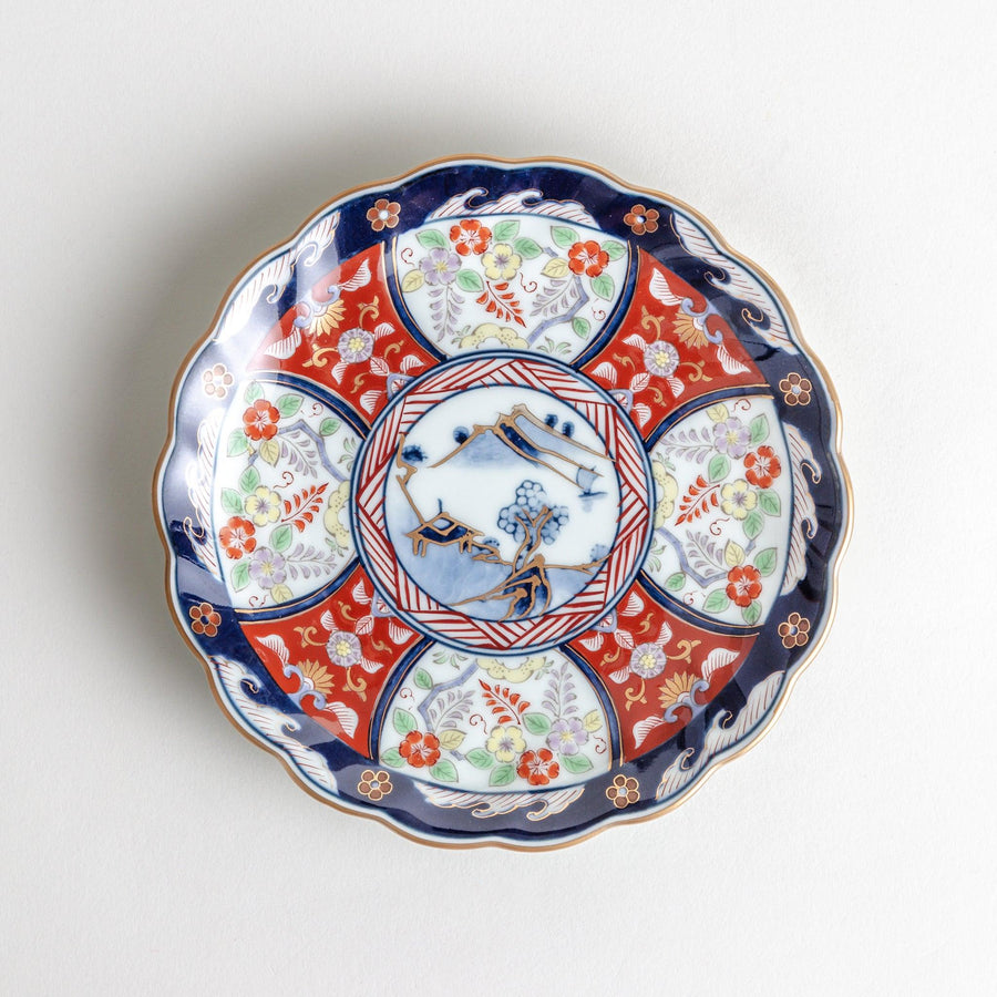 A smaller scalloped porcelain bowl with Imari-style decoration, showcasing a central landscape and vibrant red and blue motifs around the rim.