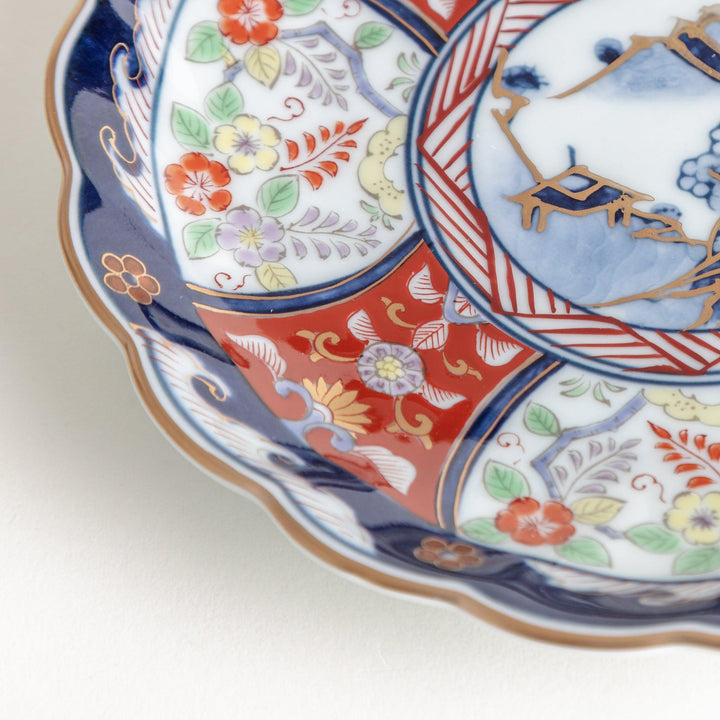 A smaller scalloped porcelain bowl with Imari-style decoration, showcasing a central landscape and vibrant red and blue motifs around the rim.