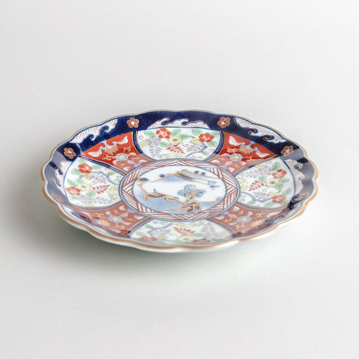 A smaller scalloped porcelain bowl with Imari-style decoration, showcasing a central landscape and vibrant red and blue motifs around the rim.