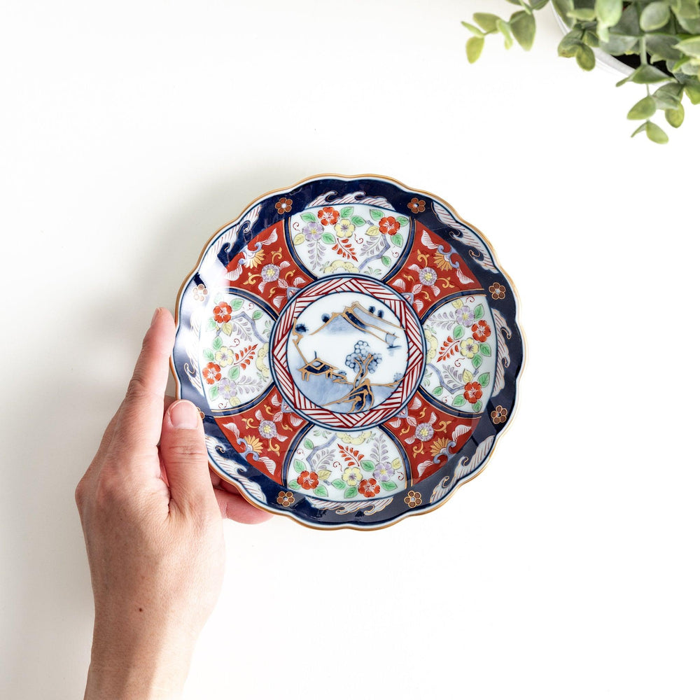 A smaller scalloped porcelain bowl with Imari-style decoration, showcasing a central landscape and vibrant red and blue motifs around the rim.