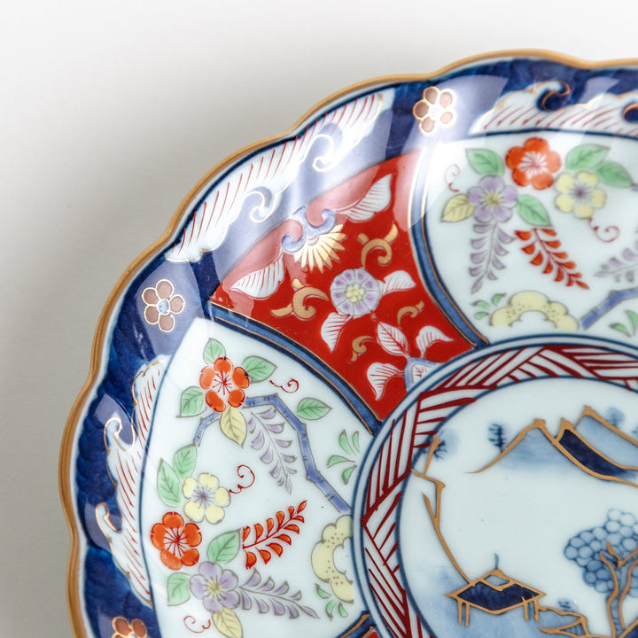 A scalloped porcelain bowl featuring an intricate Imari-style design with alternating red and blue patterns and a central landscape scene.