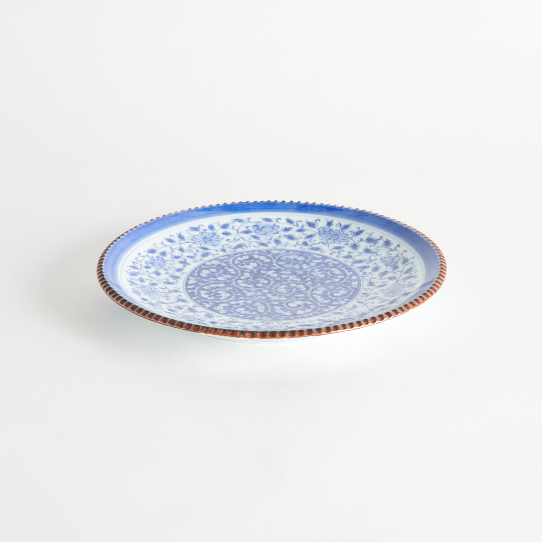 A ceramic plate with an intricate blue floral design centered around a bold circular pattern, framed by a copper-colored textured rim.