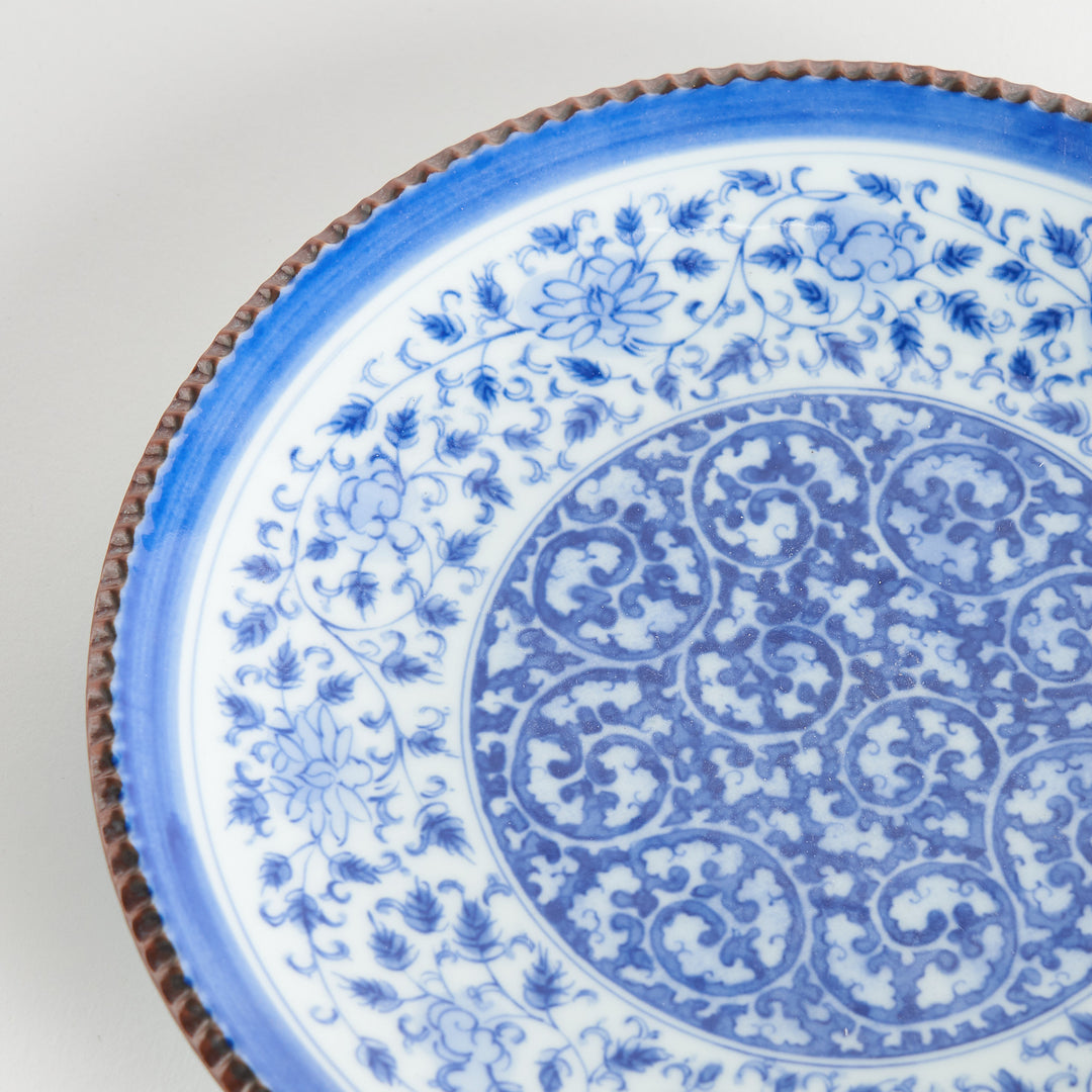 A ceramic plate with an intricate blue floral design centered around a bold circular pattern, framed by a copper-colored textured rim.