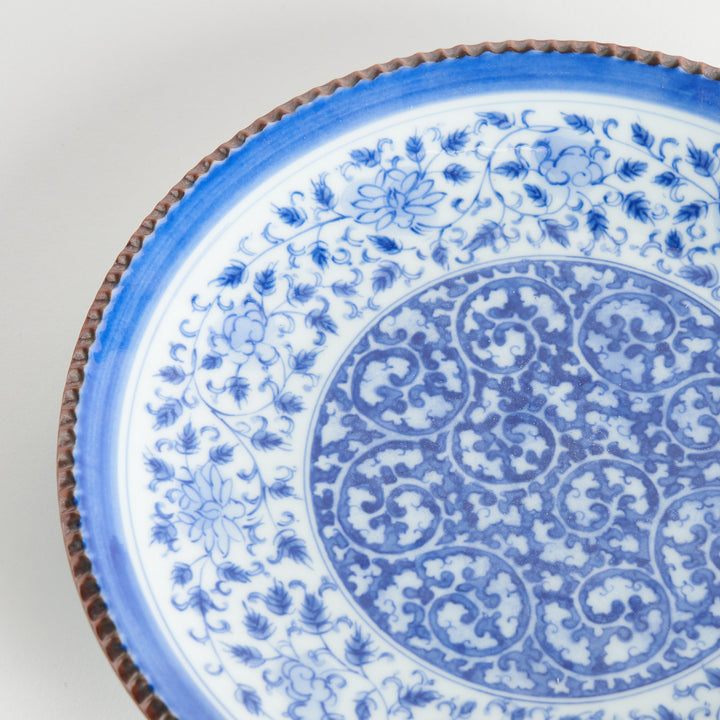 A ceramic plate with an intricate blue floral design centered around a bold circular pattern, framed by a copper-colored textured rim.