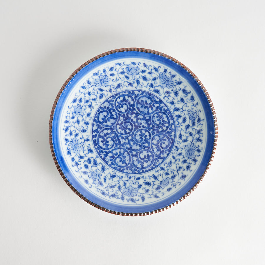 A ceramic plate with an intricate blue floral design centered around a bold circular pattern, framed by a copper-colored textured rim.