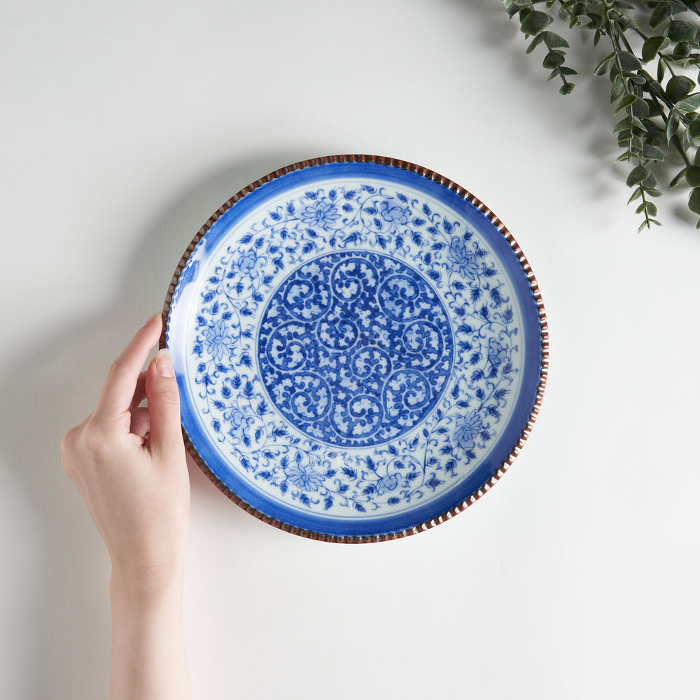 A ceramic plate with an intricate blue floral design centered around a bold circular pattern, framed by a copper-colored textured rim.