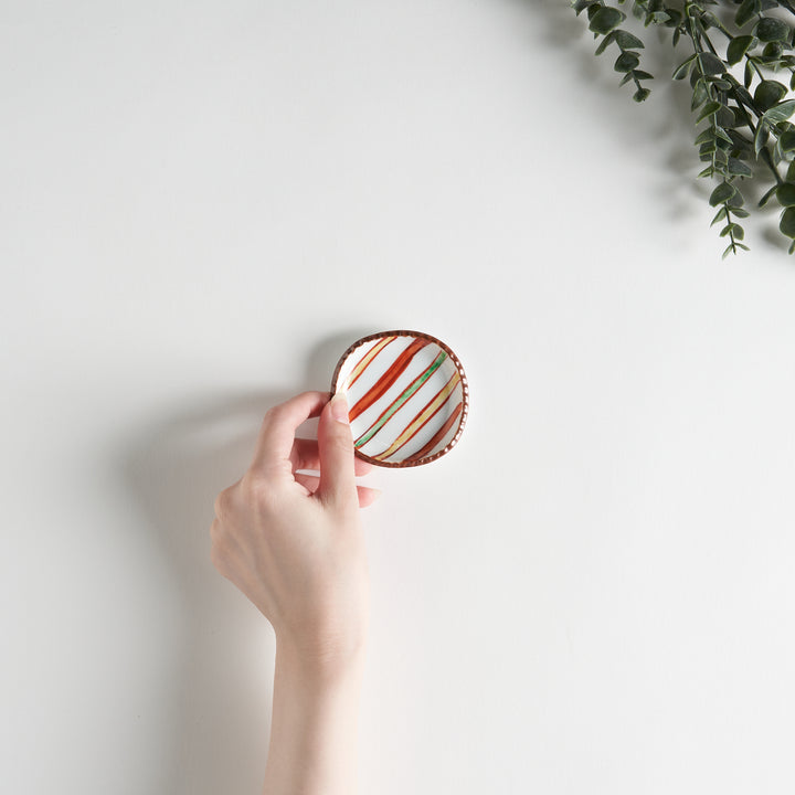 A hand holding a colorful striped sauce dish above a plain white surface, showcasing its vibrant design.