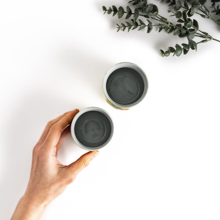 Hand holding a small teacup, next to another teacup, with eucalyptus leaves in the background.