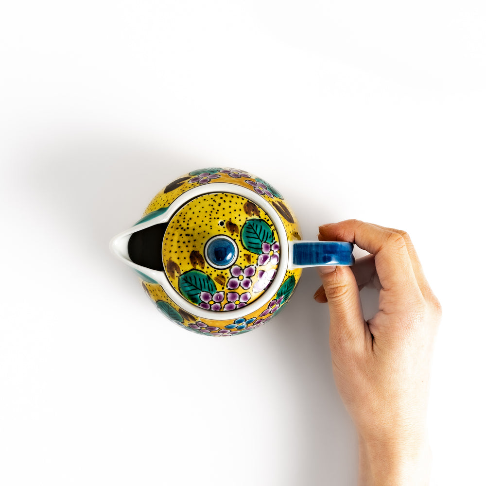 Hand holding the handle of a vibrant yellow teapot with a colorful hydrangea pattern, viewed from above.