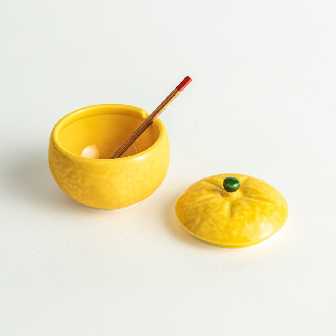 A small ceramic container shaped like a yuzu fruit with a lid and a spoon, bright yellow color.