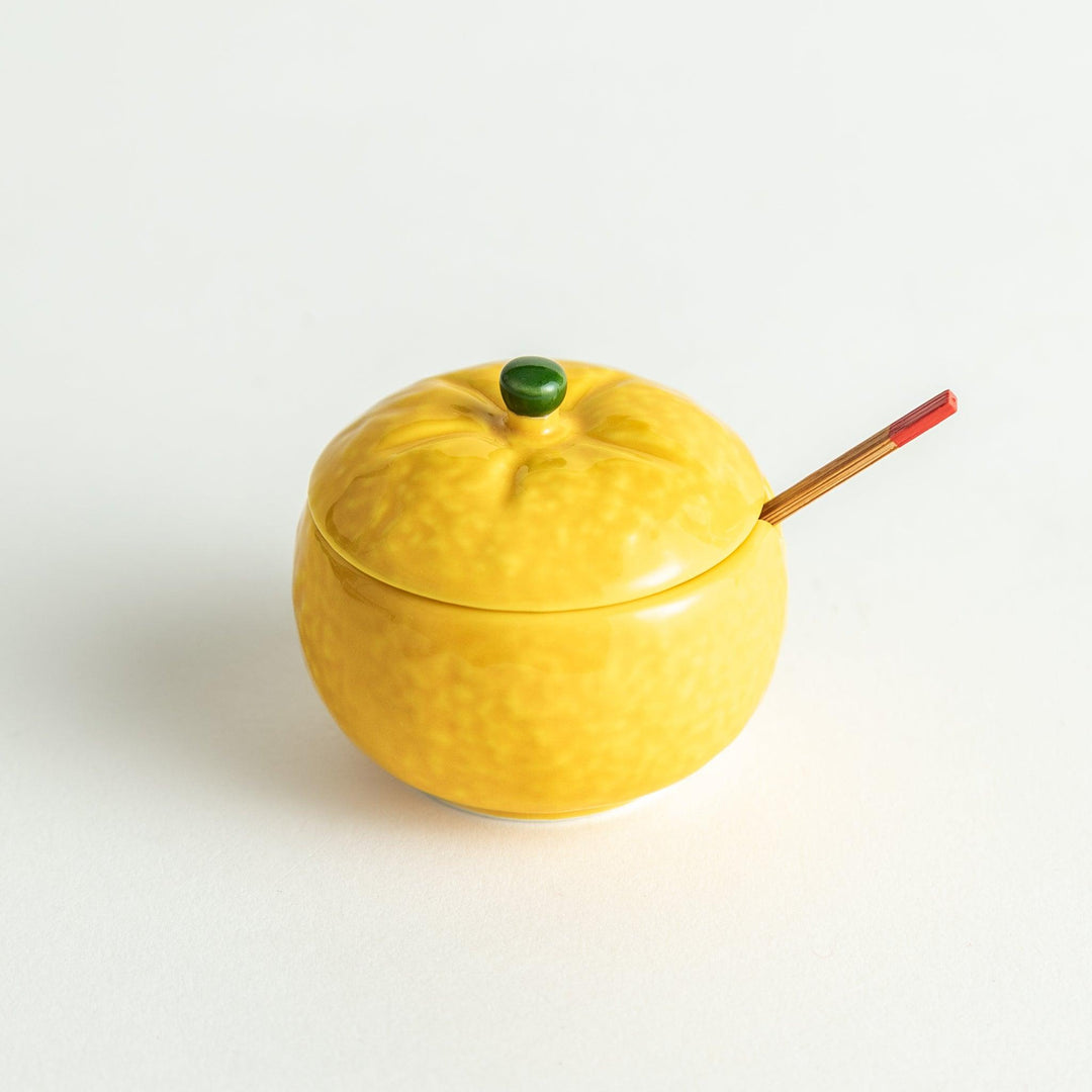 A small ceramic container shaped like a yuzu fruit with a lid and a spoon, bright yellow color.