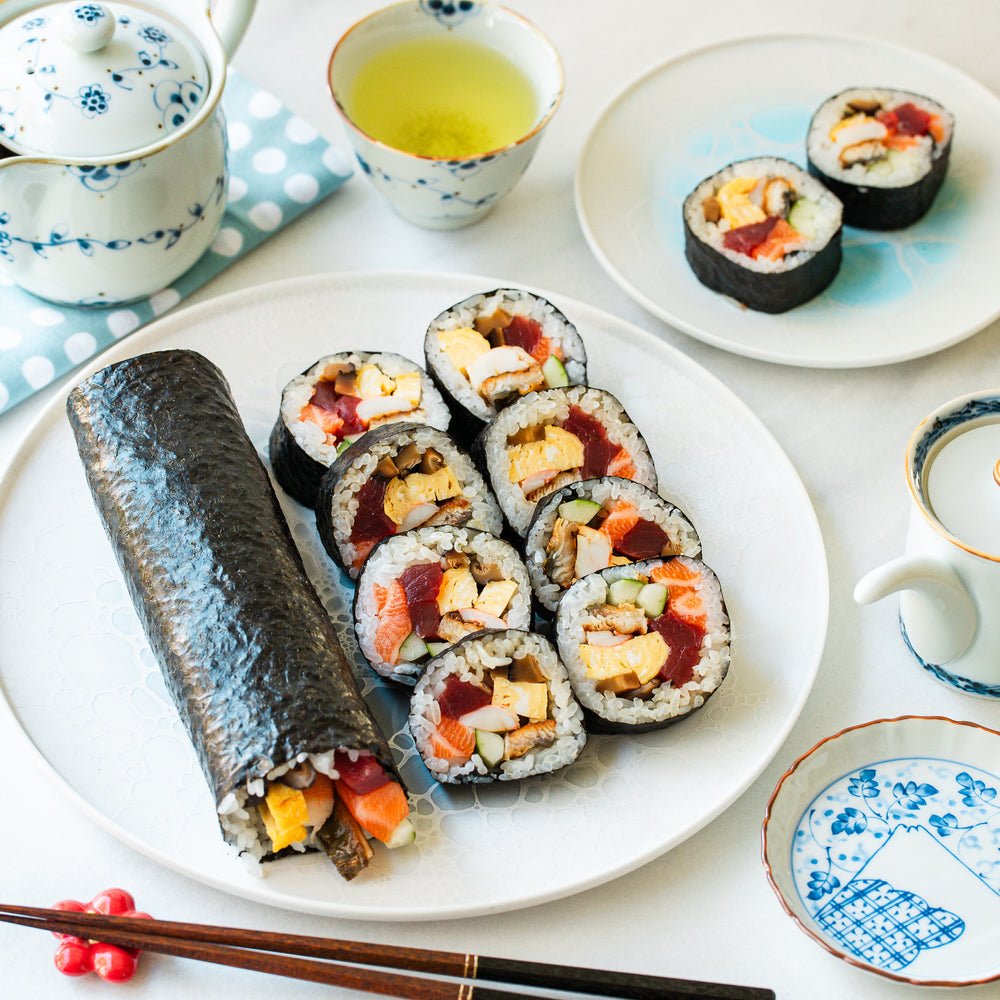 Sliced sushi rolls with tuna, egg, and vegetables on a plate, accompanied by green tea and a whole roll on the side.