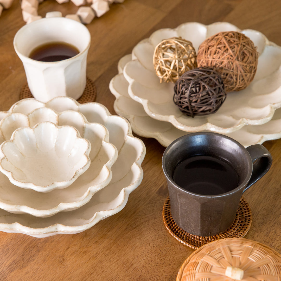 Two ceramic mugs, one rustic black and one white, sit on woven coasters amid natural-toned petal plates and decorative woven spheres, creating a cozy atmosphere.