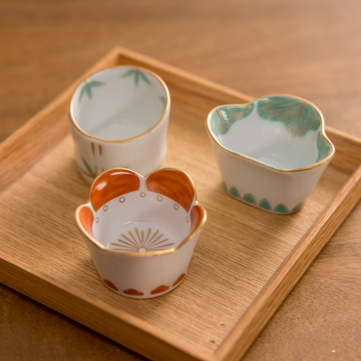 Three small bowls with floral designs: one with bamboo design, one with plum design, and one with pine design, arranged on a wooden tray.