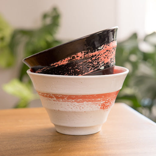 Two beautifully crafted ceramic bowls, one black with a red band and the other white with a textured orange stripe, stacked elegantly.