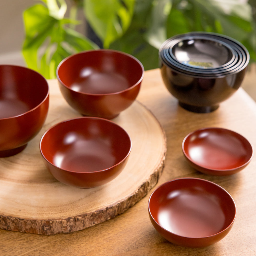 A beautifully set of nested bowls featuring a large five-piece oryoki set in a warm red finish, complemented by a sleek black bowl stack. The bowls are displayed on a natural wooden surface with soft greenery in the background.