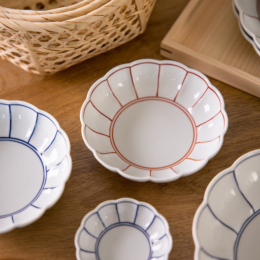 A collection of elegant ceramic bowls in various sizes, featuring petal-shaped designs with red and blue accents, displayed on a wooden table alongside a woven basket.