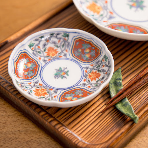 A vibrant crimson floral sauce dish rests on a wooden tray, adorned with intricate floral designs in red, blue, and green. Two chopsticks sit beside the dish, enhancing the presentation with a touch of elegance.