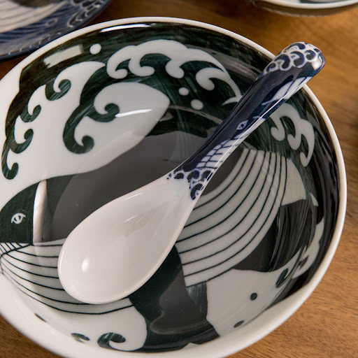 A white spoon with a blue whale design resting in a bowl, featuring a wave-like pattern in dark blue and white.