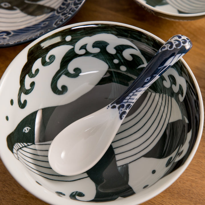 A white spoon with a blue whale design resting in a bowl, featuring a wave-like pattern in dark blue and white.