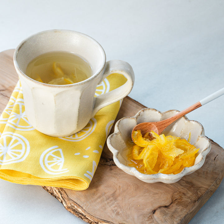 Tea in a rustic mug with yuzu slices, paired with yuzu syrup in a small bowl, and a wooden spoon. Yellow lemon-printed napkin adds a vibrant touch.