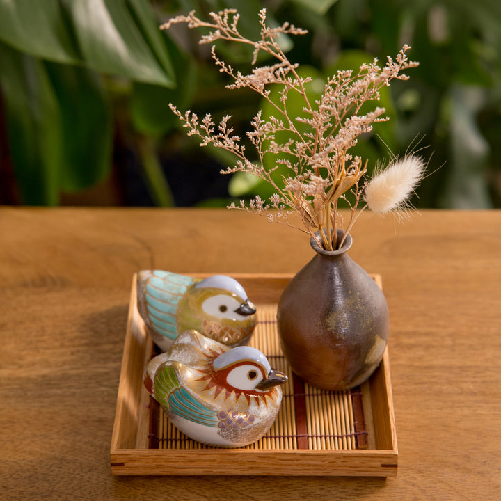 Two colorful bird-shaped bowls sit on a bamboo mat tray beside a small vase holding dried flowers, enhancing the cozy decor of the table setting.