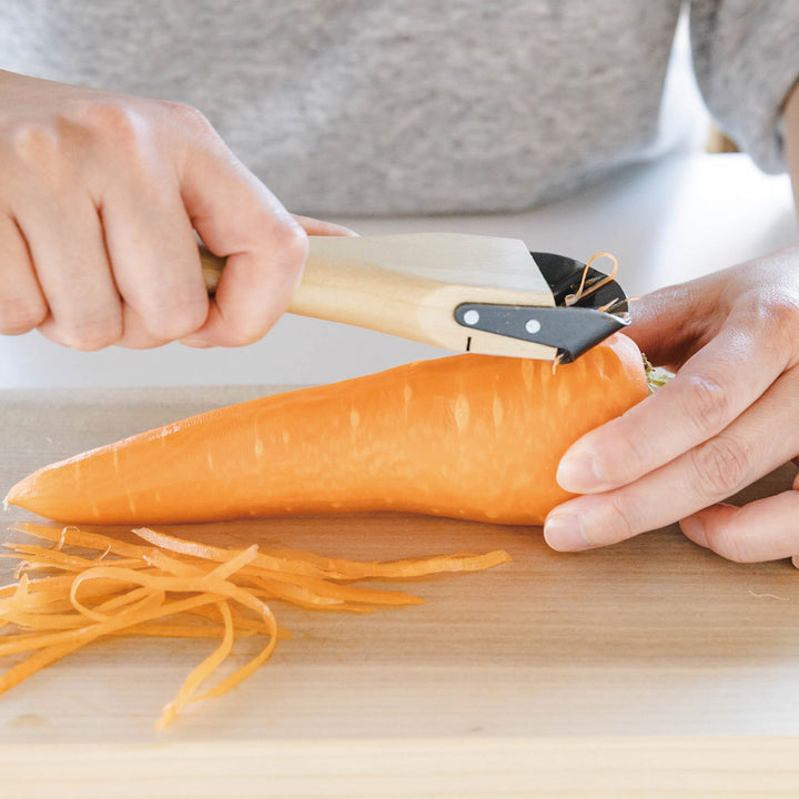 A Wood Handle Julienne Peeler, a kitchen tool used to create thin vegetable strips from a carrot, perfect for salads and garnishes.