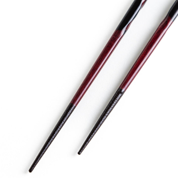 A pair of twisted wooden chopsticks in a dark red and black color gradient.